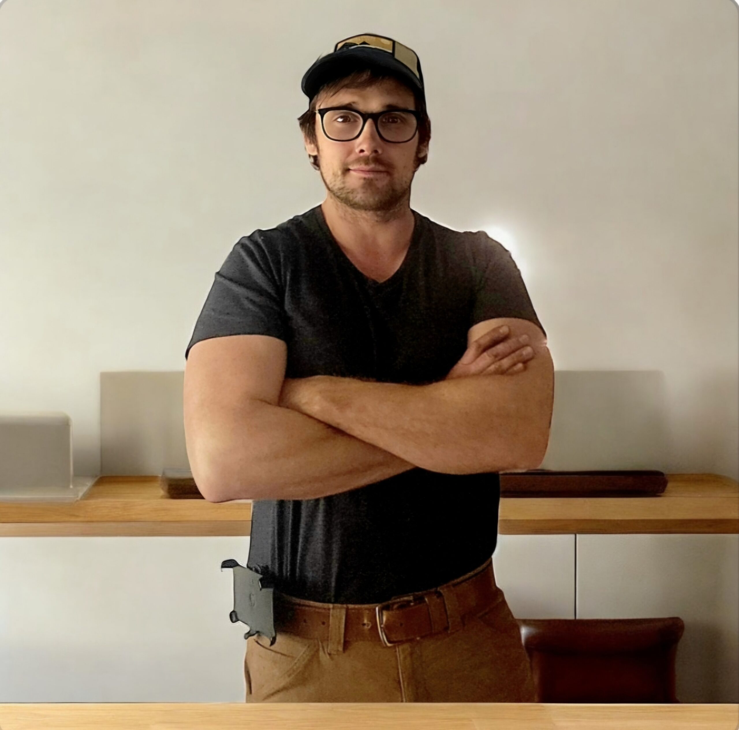A man wearing glasses, a dark T-shirt, and a cap stands with arms crossed in front of a wooden counter in a minimalist room.