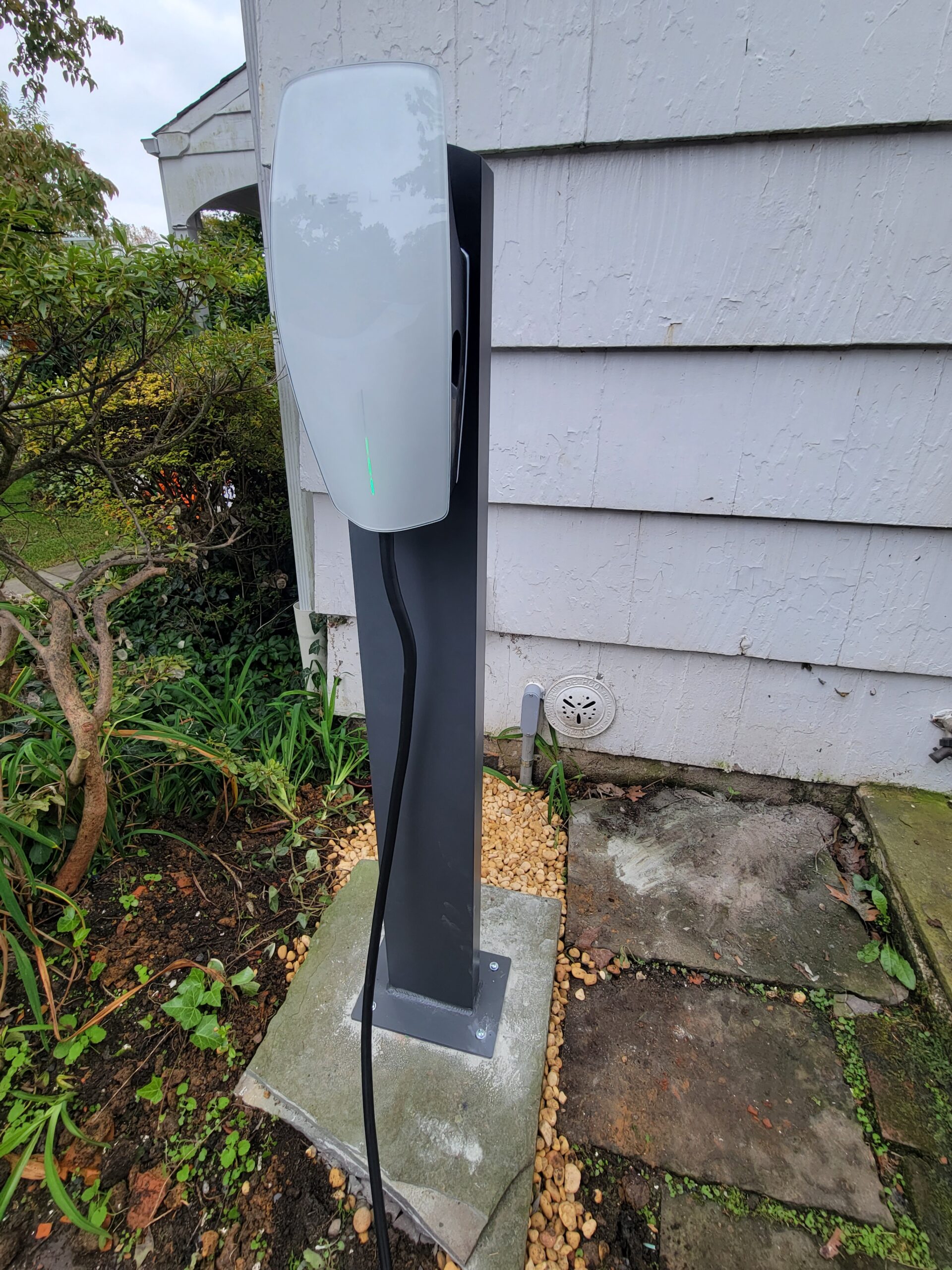 An electric vehicle charging station is installed on a concrete base next to the exterior wall of a house, surrounded by plants and stone pavers.