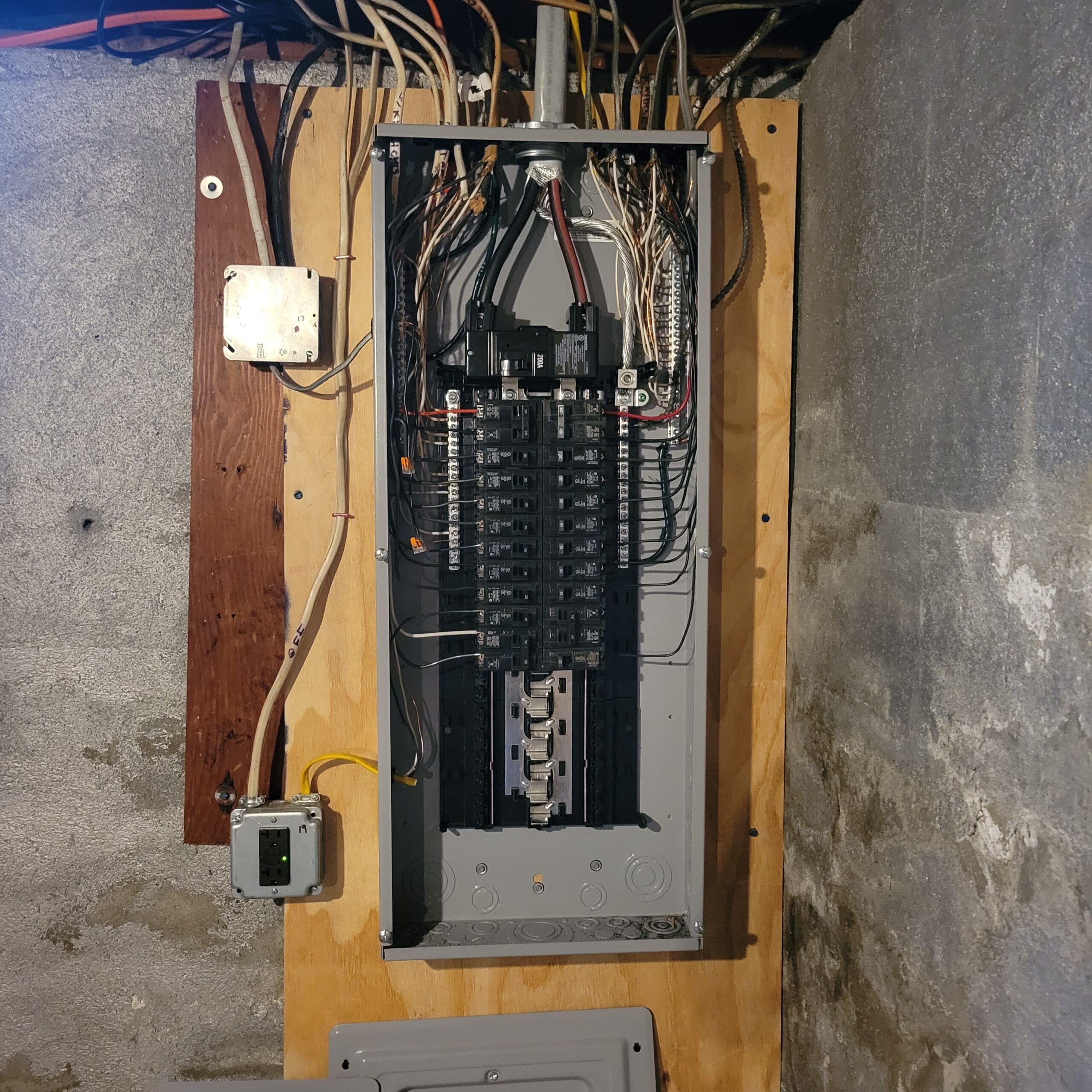 An open electrical panel with multiple circuit breakers, wiring, and cables mounted on a plywood board against a basement wall.