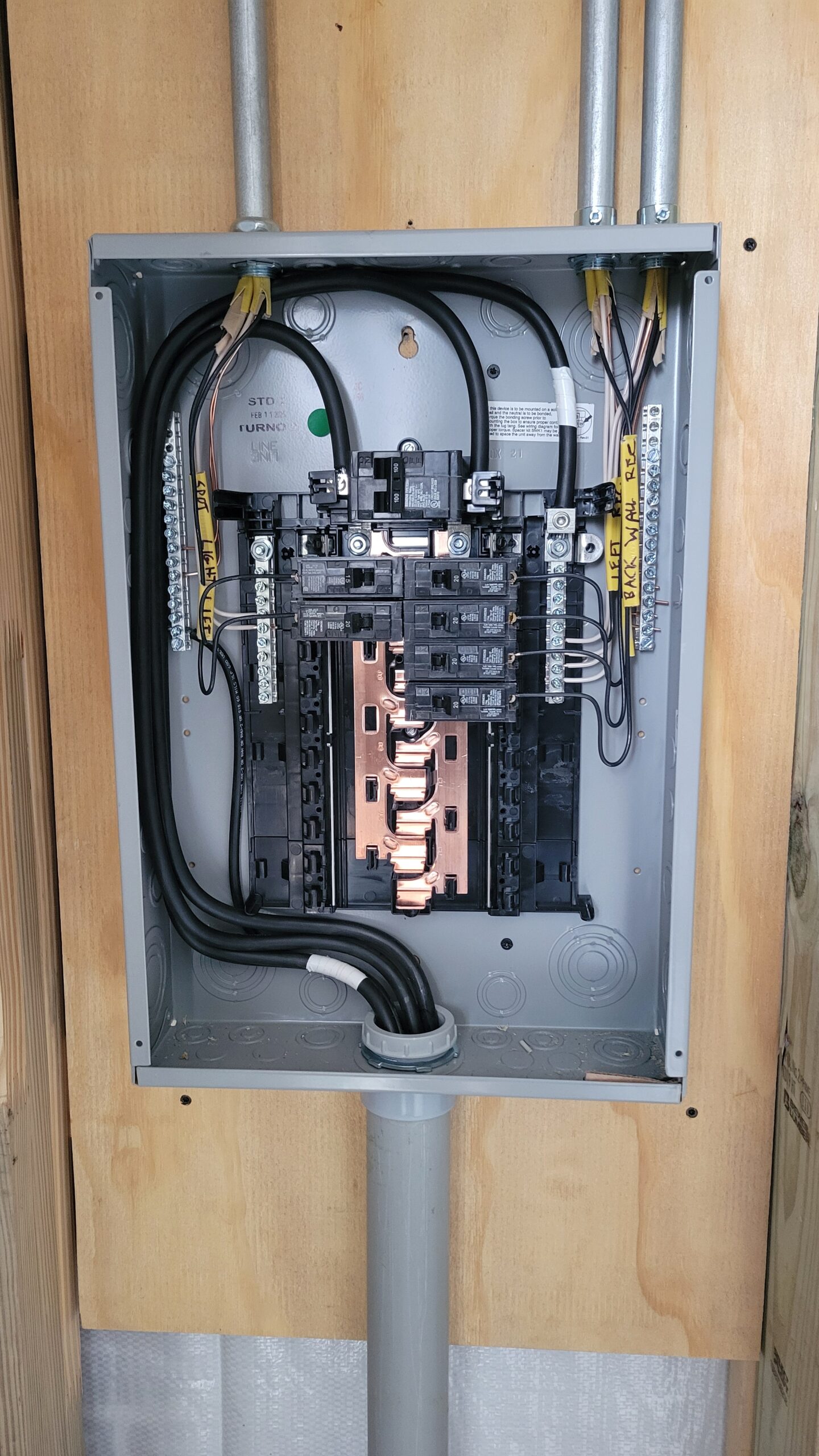 An open electrical panel with exposed circuit breakers, labeled wires, and conduit pipes mounted on a plywood wall.