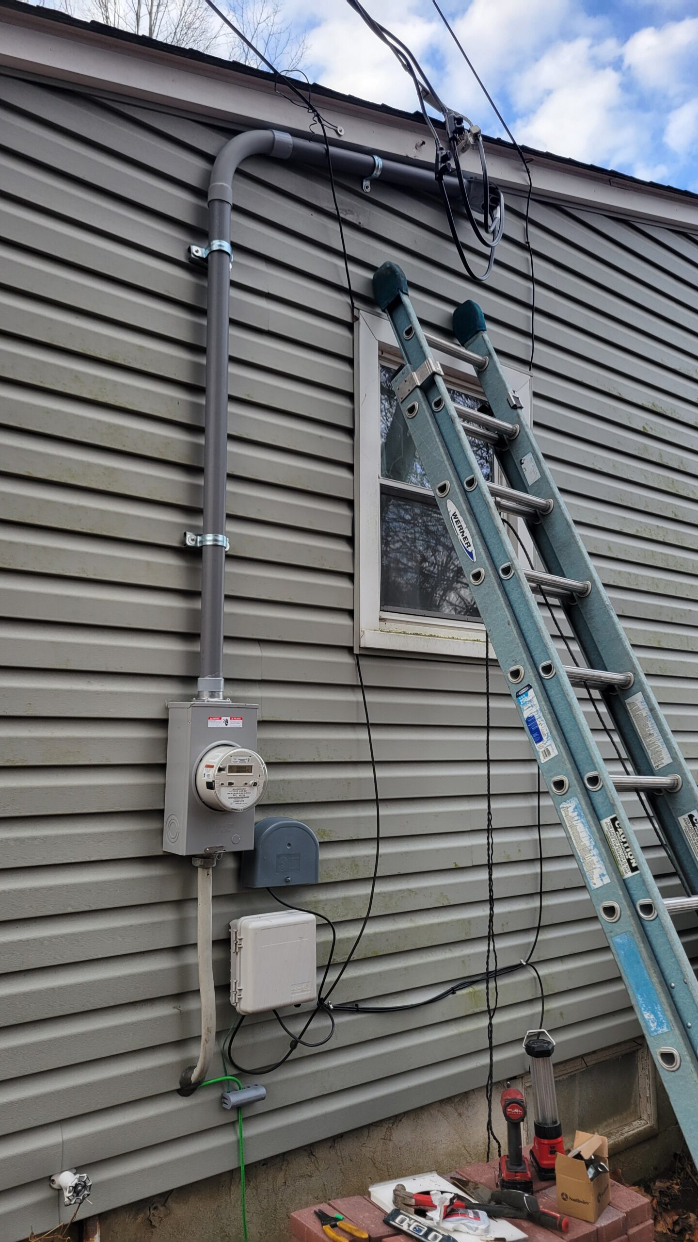 A ladder leans against a house wall near a window; electrical meter, conduit, cables, and tools are visible, indicating ongoing electrical work.