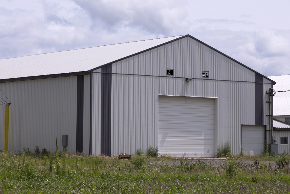 A large white metal warehouse building with a closed garage door sits on a grassy lot under a cloudy sky.