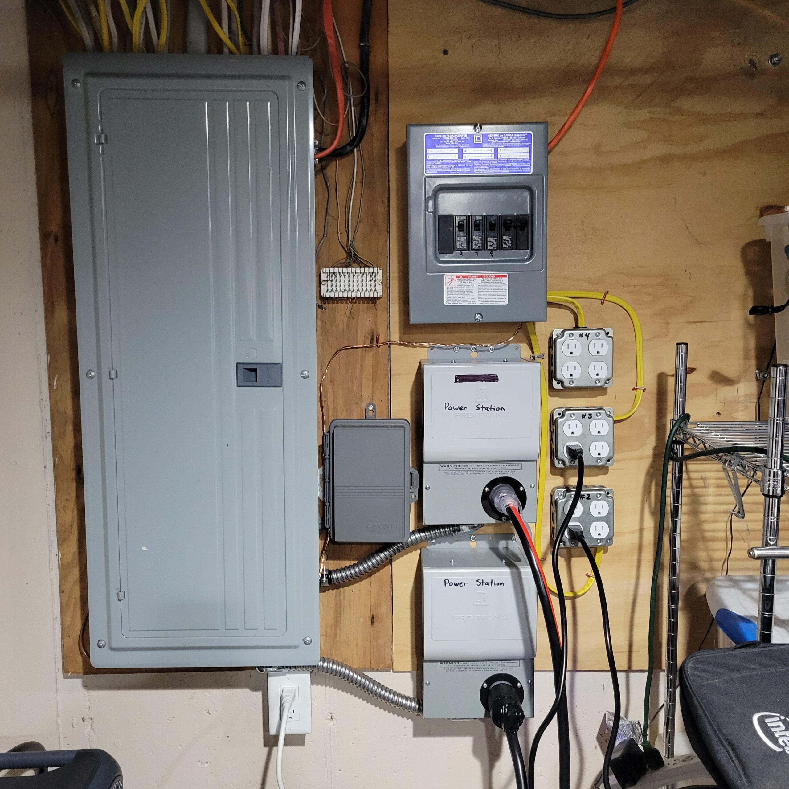 Electrical panel and subpanel mounted on a plywood wall, with labeled power stations, multiple outlets, wiring, and conduit visible.