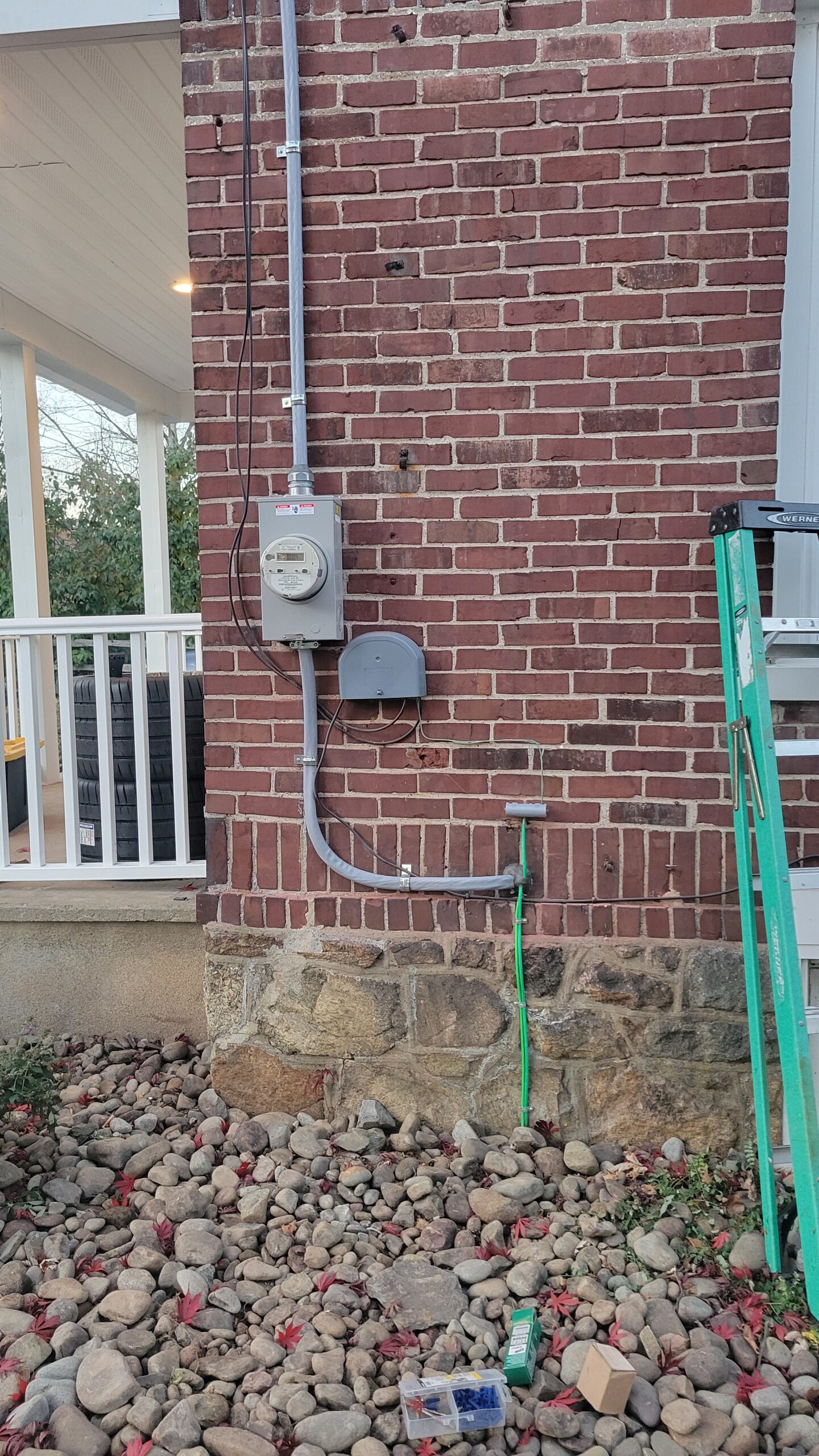 Electrical meter and junction box mounted on a brick wall, with conduit running down to an outlet. A green garden hose, a green ladder, and a small toolbox are visible nearby.